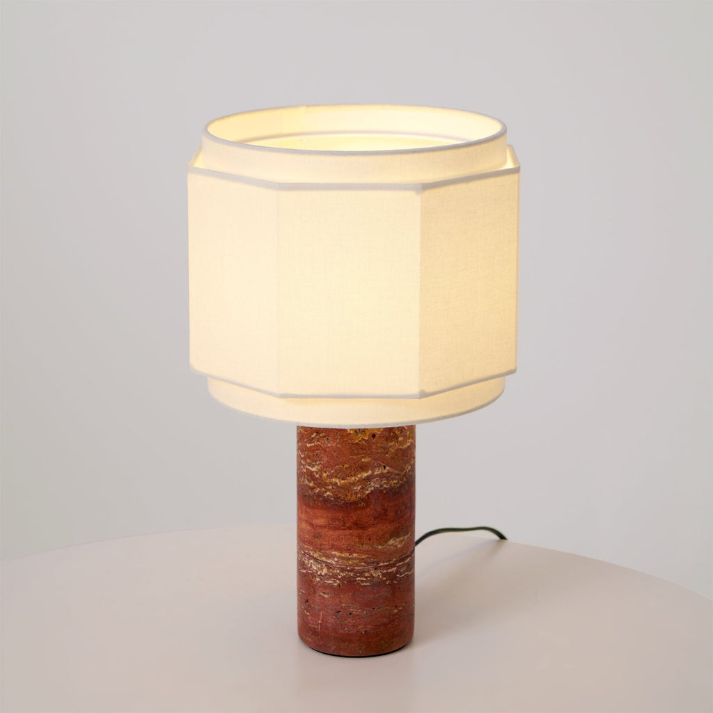Set against a bare backdrop, this Clérachic table lamp's red travertine column rises from a plain white surface.