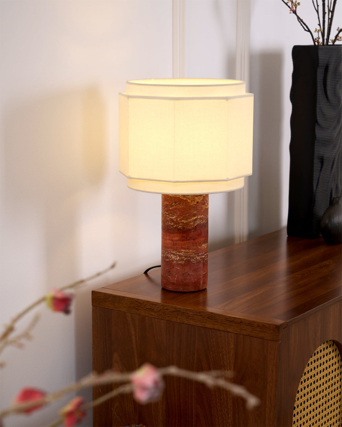 This Clérachic table lamp glows on a wood cabinet edge, a woven cane chair back and blossom sprigs blurred behind it.