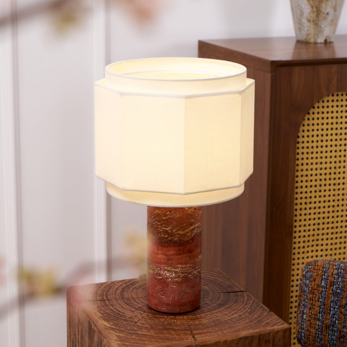 The red travertine table lamp from Clérachic lit on a wood plinth, a tall vase and cane chair softly blurred behind.
