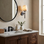 Next to a round black-framed mirror, a matte black Clérachic wall light sits above a walnut vanity with a small vase.