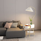 This Clérachic floor lamp's white built-in table sits beside a grey sofa, its pleated shade glowing on a curved brass stem.
