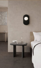 Mounted above a dark bedside table set with a glass carafe and bowls, this Clérachic wall sconce lights a calm bedroom corner.