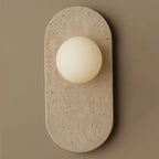 Cream travertine with natural surface pitting forms the oval body of this Clérachic wall sconce, shown close against a taupe wall.