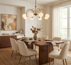 Cream curved-back chairs surround a wood table lit by this brass Clérachic chandelier, with a framed painting nearby.