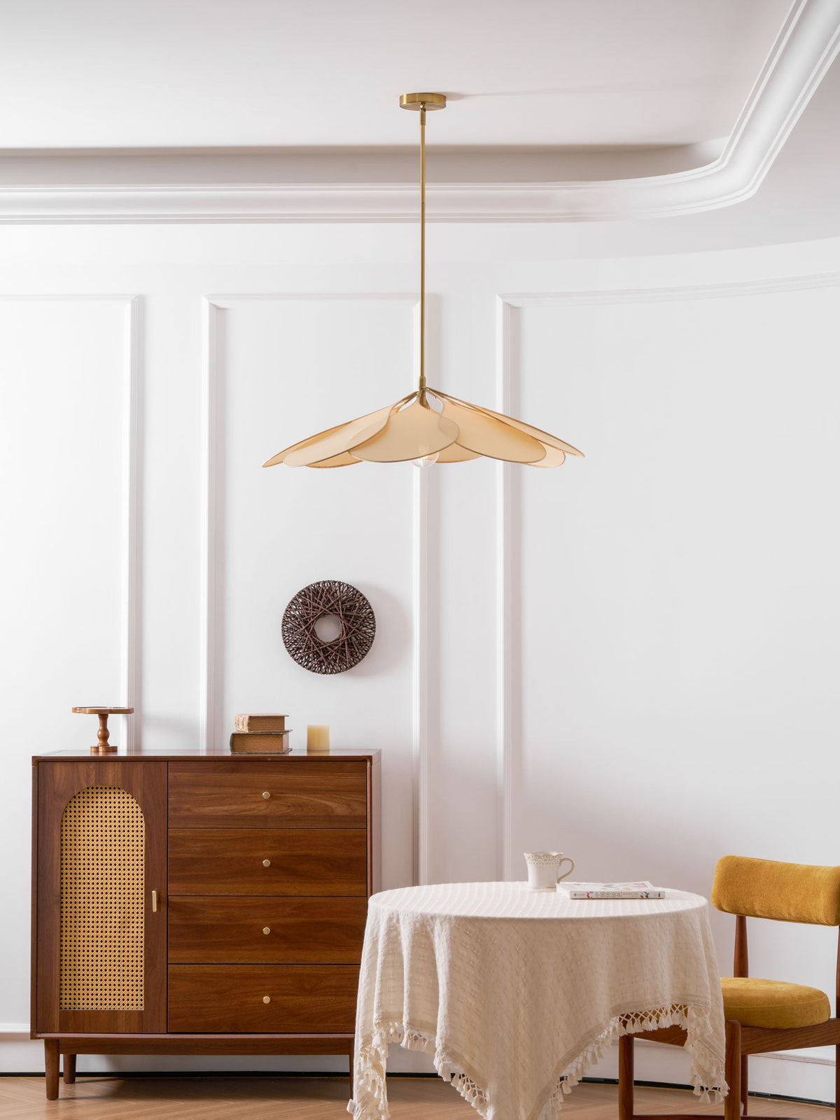 This Clérachic pendant lamp in beige petals hangs near a walnut sideboard and a round table set with a jug and mustard chair.