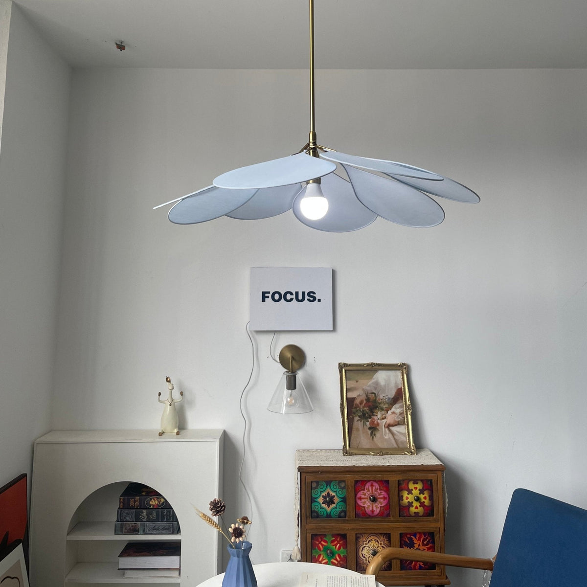 This Clérachic pendant lamp in pale blue petals hangs above a white console, with a framed picture and blue chair nearby.