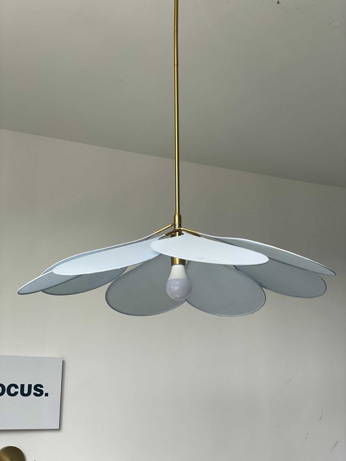 This Clérachic pendant lamp in pale blue petals hangs from a long brass stem beneath a sloped white ceiling, its bulb visible.
