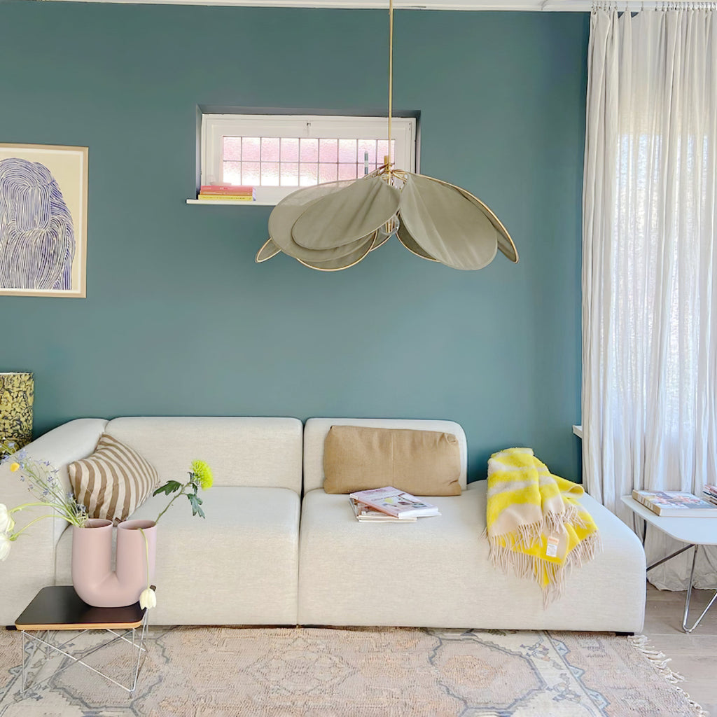 This Clérachic pendant lamp in a khaki-toned version hangs against a teal wall, above a beige sofa with striped cushions.