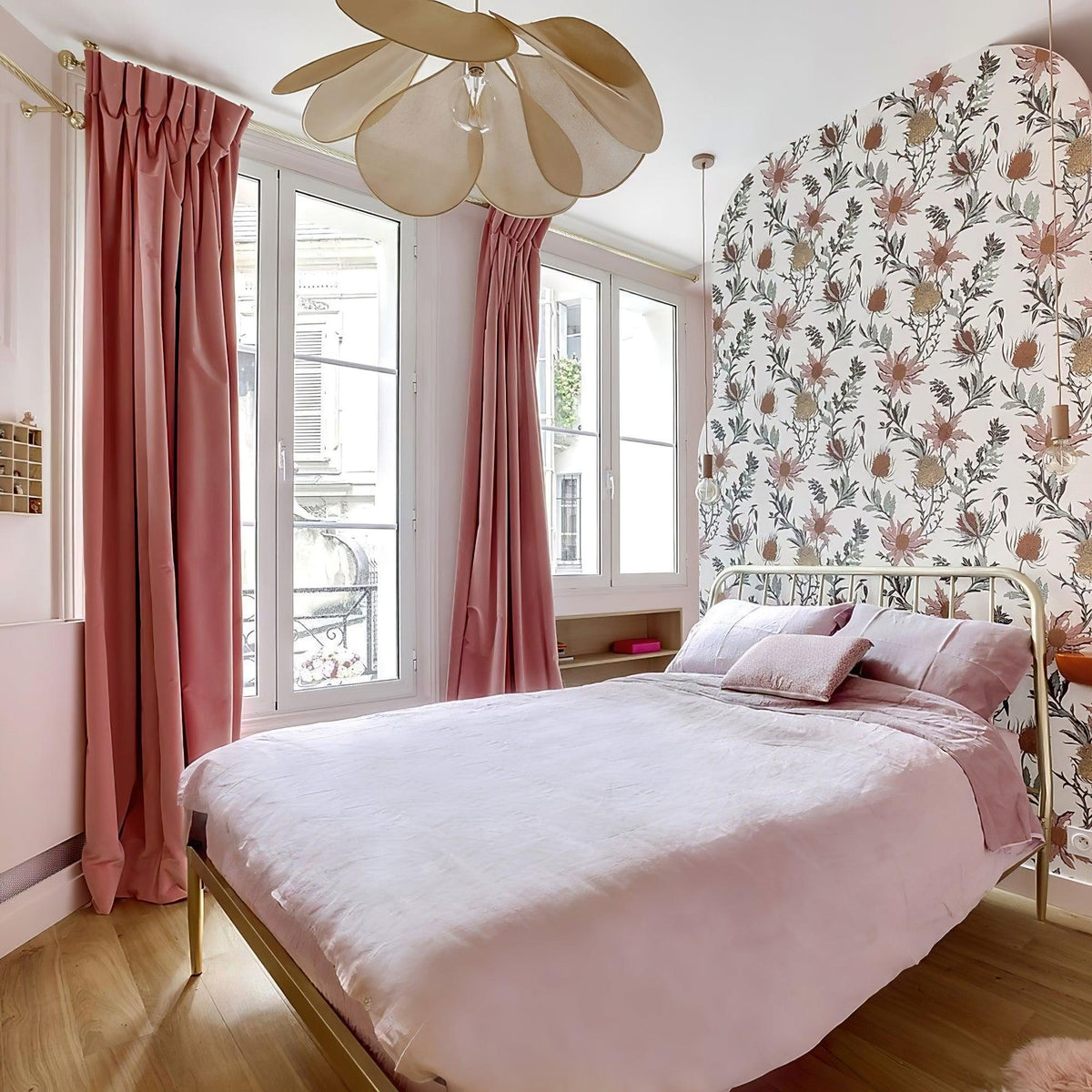 This Clérachic pendant lamp in cream petals hangs above a brass bed dressed in pink linens, beside a floral-papered wall.