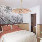This Clérachic pendant lamp in tan petals hangs above a bed dressed in blush pink linens, beside a black-and-white palm-print wall.