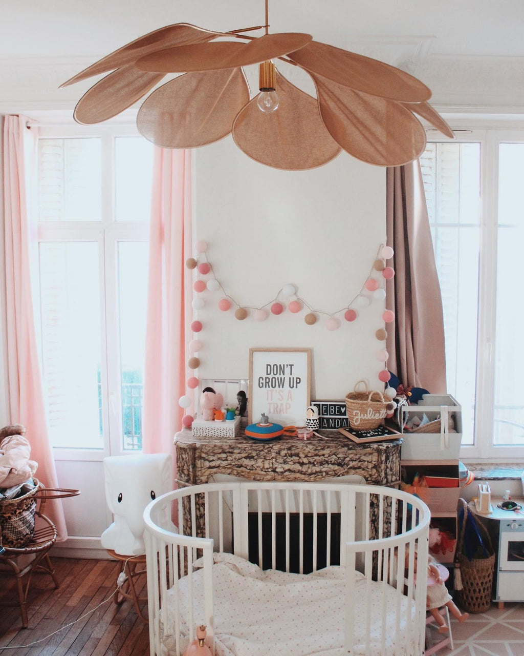This Clérachic pendant lamp in copper-toned petals hangs above a round crib in a nursery decorated with a pastel pom-pom garland.