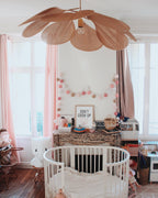 This Clérachic pendant lamp in copper-toned petals hangs above a round crib in a nursery decorated with a pastel pom-pom garland.