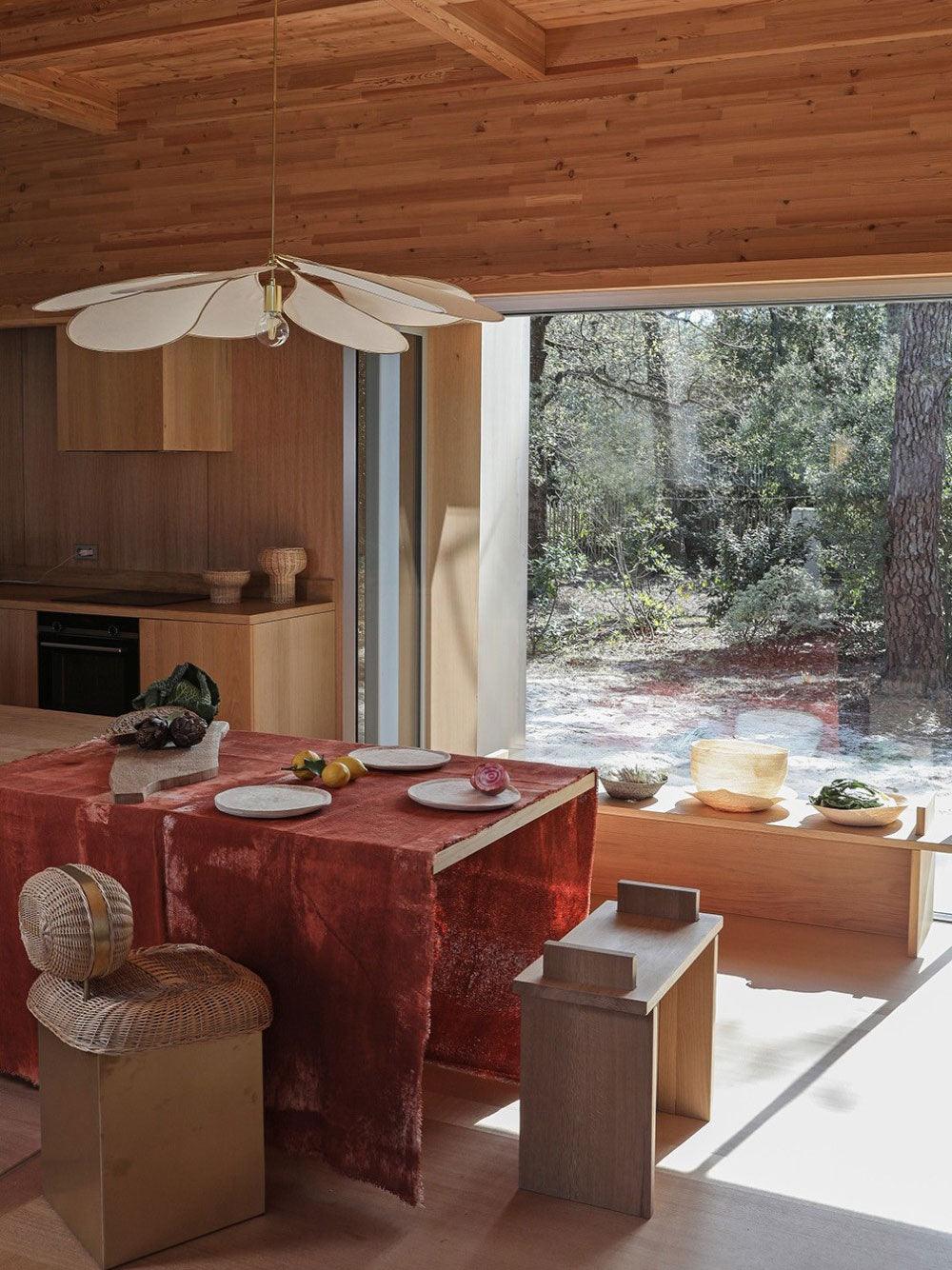 This Clérachic pendant lamp in white fabric petals hangs above a dining table dressed with a red cloth, in a timber-lined room overlooking a forest.