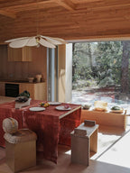 This Clérachic pendant lamp in white fabric petals hangs above a dining table dressed with a red cloth, in a timber-lined room overlooking a forest.