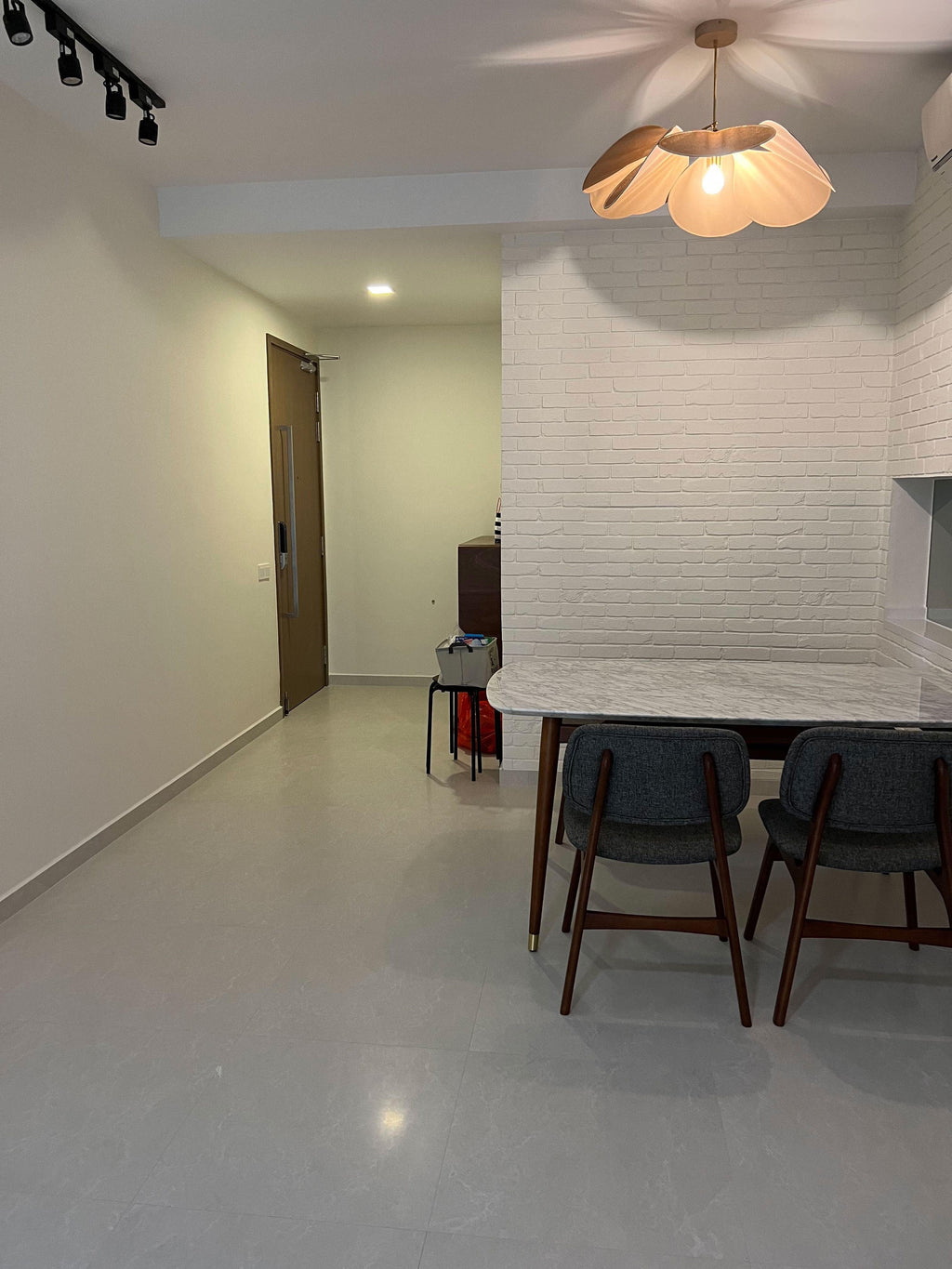 This Clérachic pendant lamp in tan petals hangs in a hallway with a white brick wall and dark dining chairs beyond.