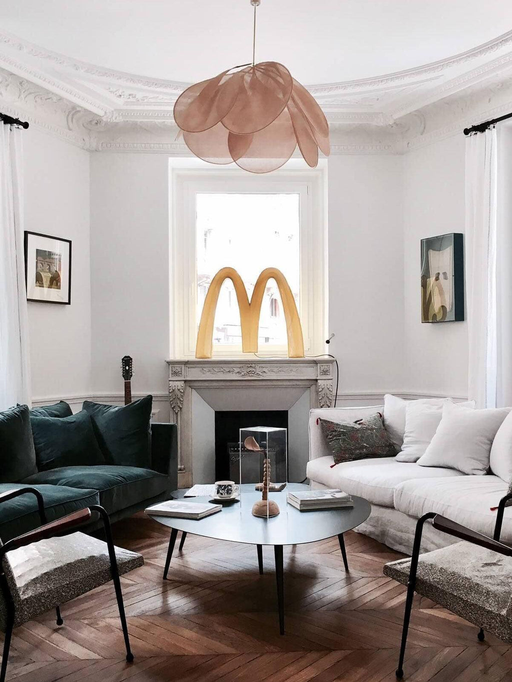 This Clérachic pendant lamp in tan petals hangs in a living room with a dark green velvet sofa and a marble fireplace.