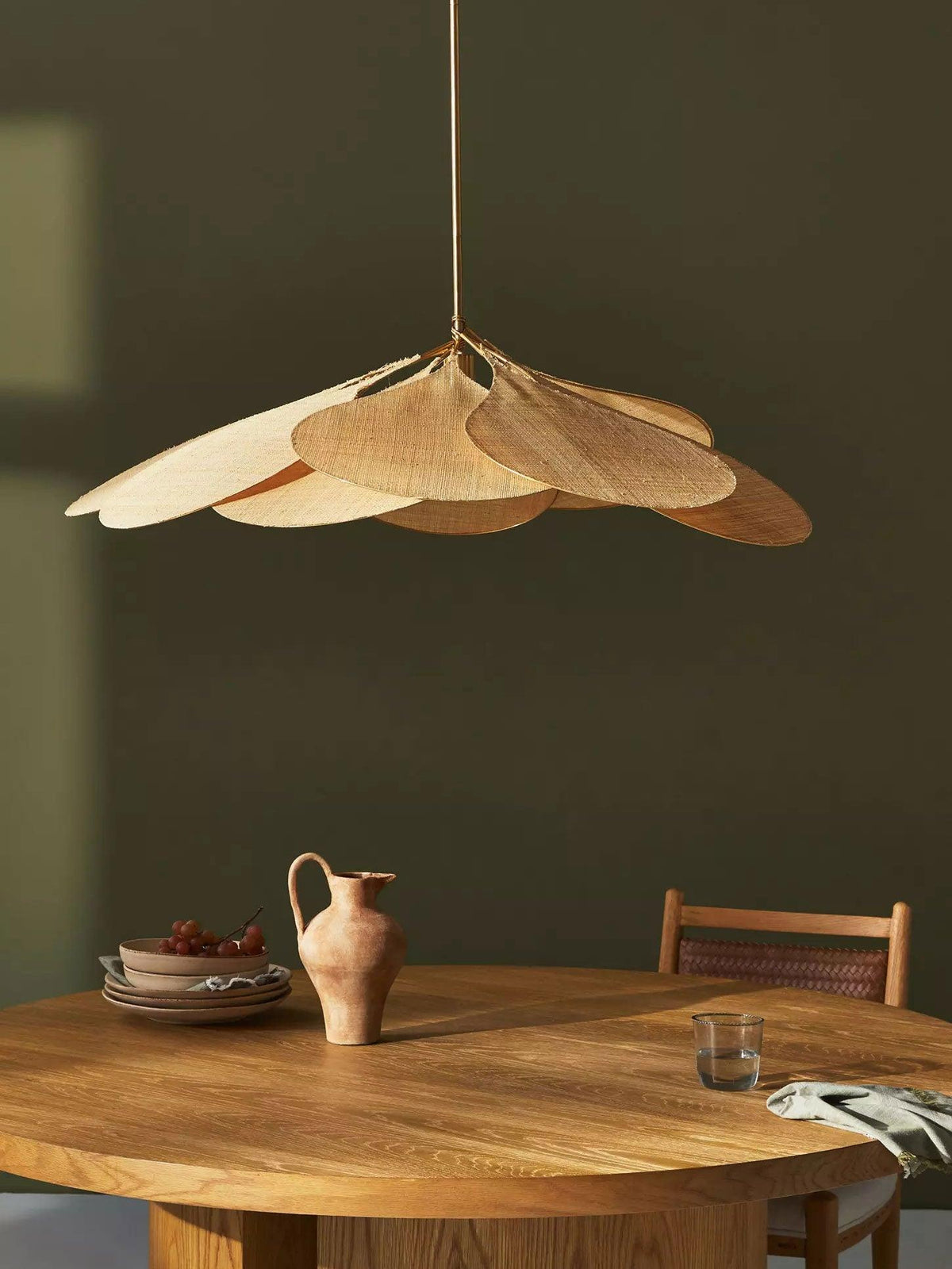 Close view of this Clérachic pendant lamp's tan petals against an olive-green wall, above a round table set with a jug and plates.