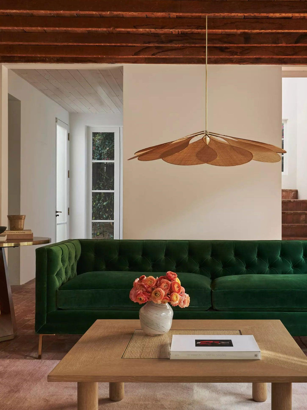 This Clérachic pendant lamp in tan petals hangs in a living room with a dark green tufted sofa and a low wooden coffee table.