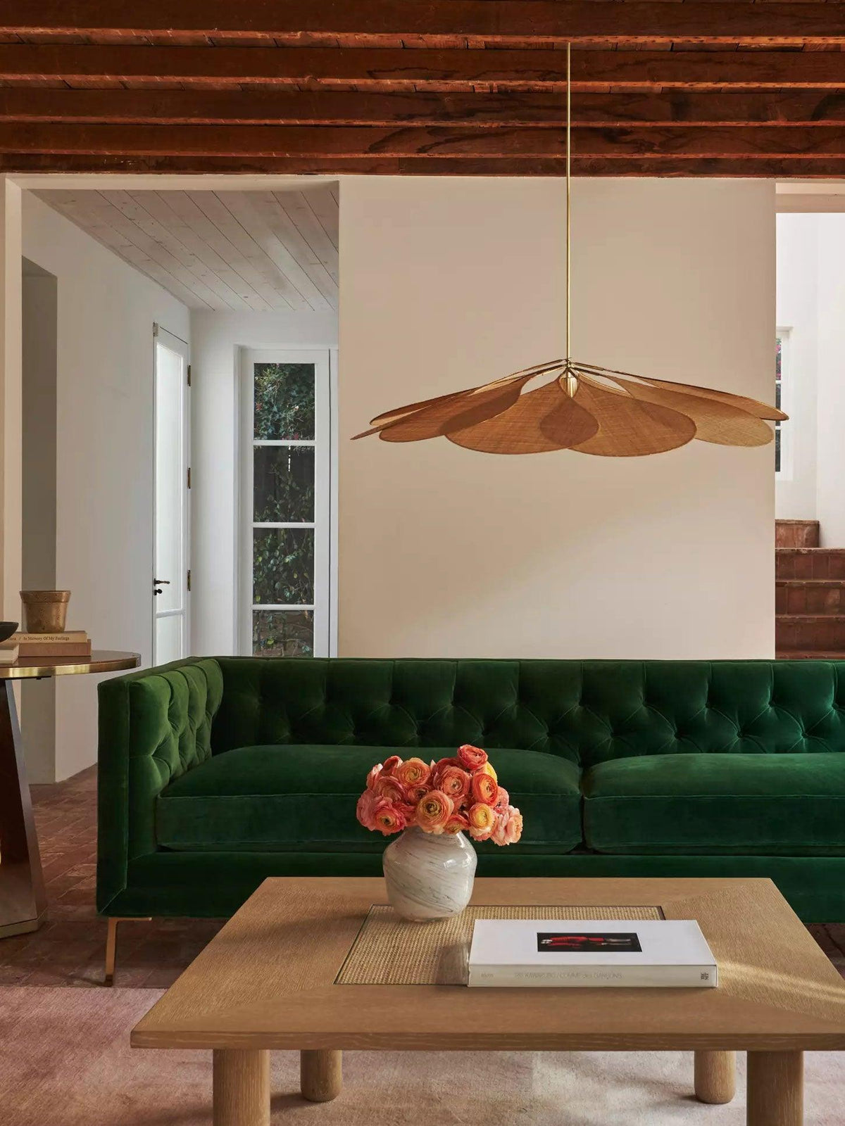 This Clérachic pendant lamp in tan petals hangs in a living room with a dark green tufted sofa and a low wooden coffee table.