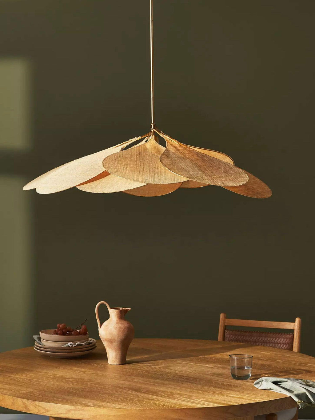 Close view of this Clérachic pendant lamp's tan petals against an olive-green wall, above a round table with a terracotta jug.
