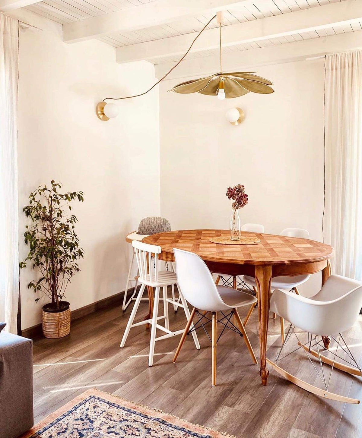 This Clérachic pendant lamp in tan petals hangs above a round wooden table with white chairs in a light-filled dining nook.