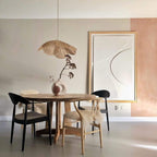 This Clérachic pendant lamp in tan petals hangs above a round wooden table with black and cane chairs, beside an abstract print.