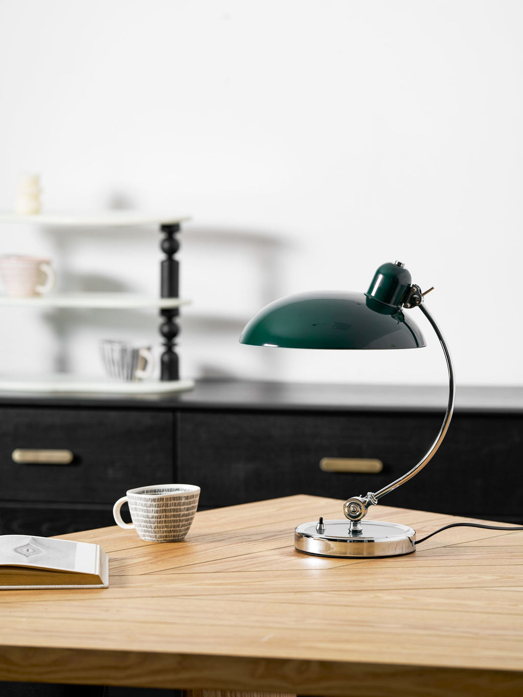 Unlit, this green Clérachic desk lamp rests on a wooden table beside a patterned cup and blurred shelving.