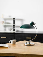 Unlit, this green Clérachic desk lamp rests on a wooden table beside a patterned cup and blurred shelving.