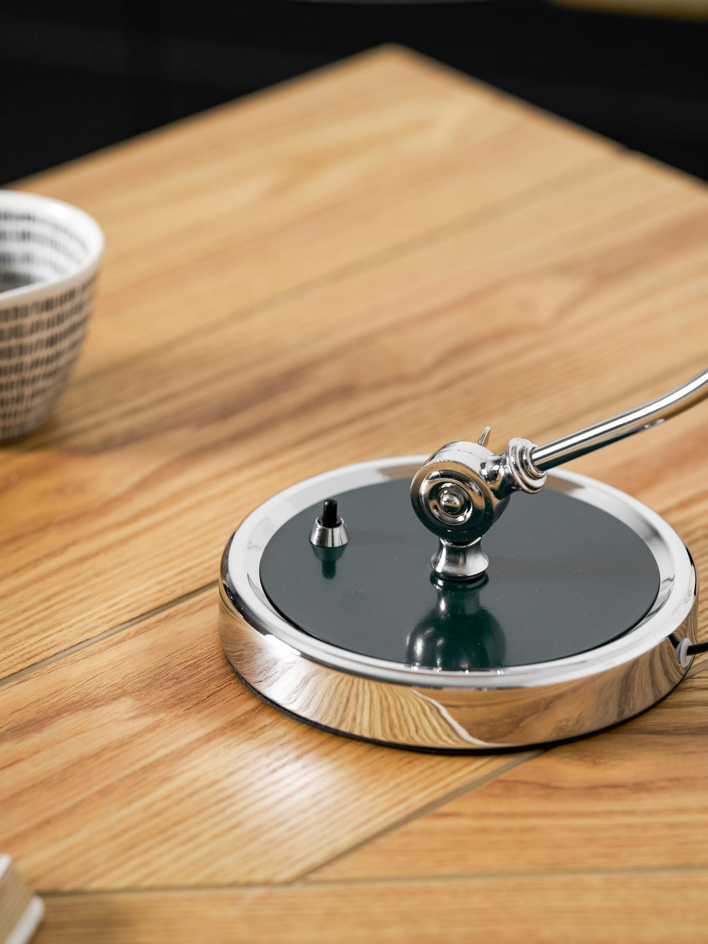 Close view of the chrome swivel joint and green base disc on this Clérachic desk lamp, set on a wood table.