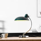 A green Clérachic desk lamp lit on a wooden table with a cup, its cord visible against blurred shelving.
