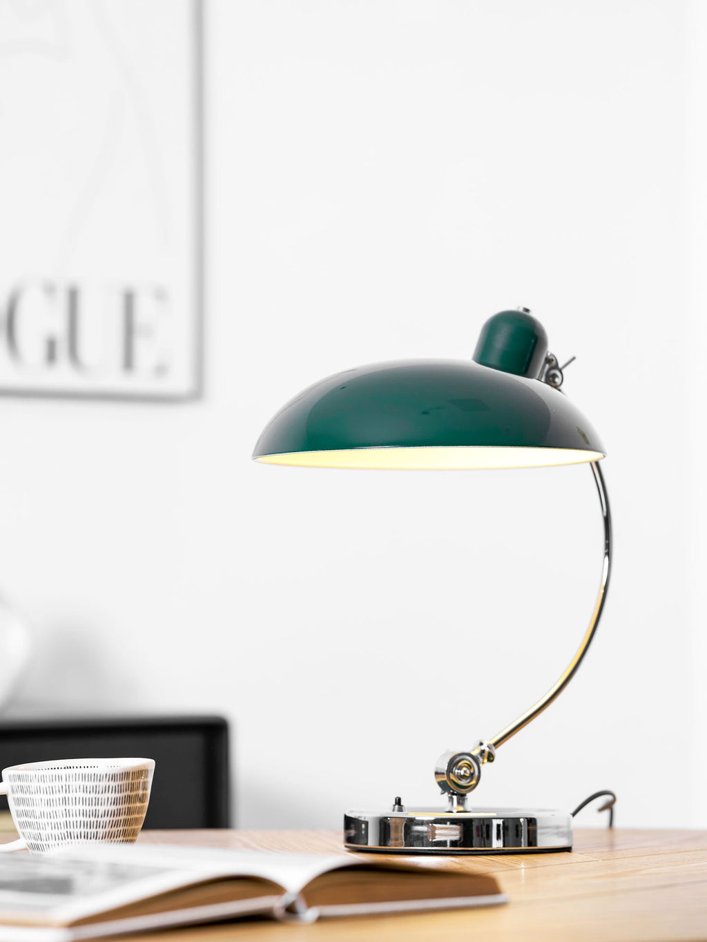 A green Clérachic desk lamp lit on a wooden table beside an open magazine and framed print.