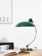 A green Clérachic desk lamp lit on a wooden table beside an open magazine and framed print.