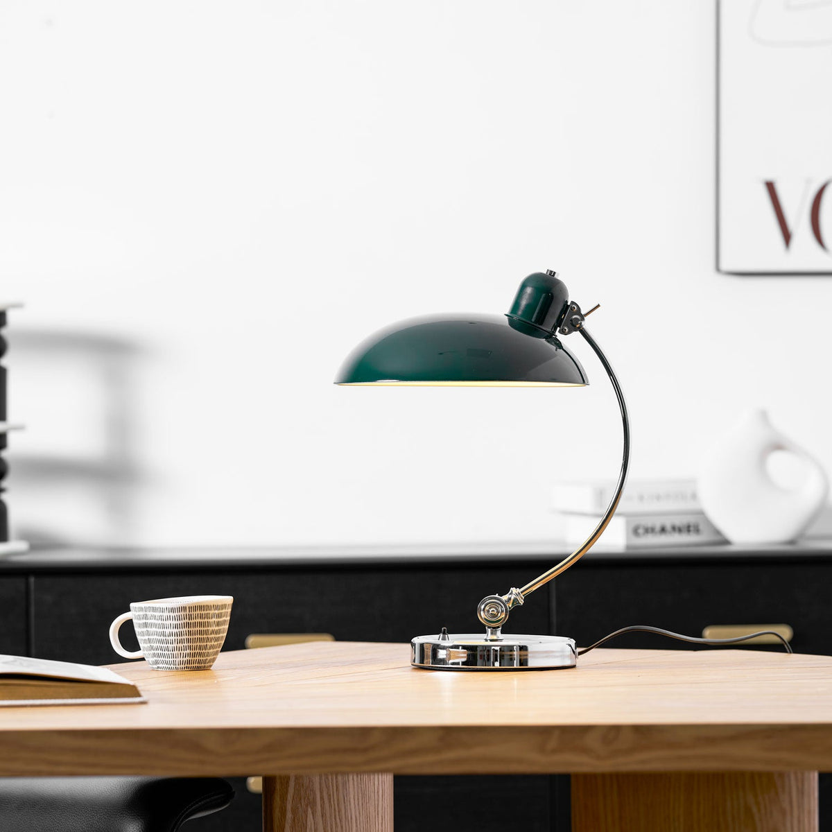 Lit on a wooden table, this green Clérachic desk lamp sits near a patterned cup with kitchen shelving blurred behind.