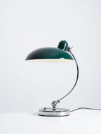 A green Clérachic desk lamp lit on a plain white backdrop, full length.
