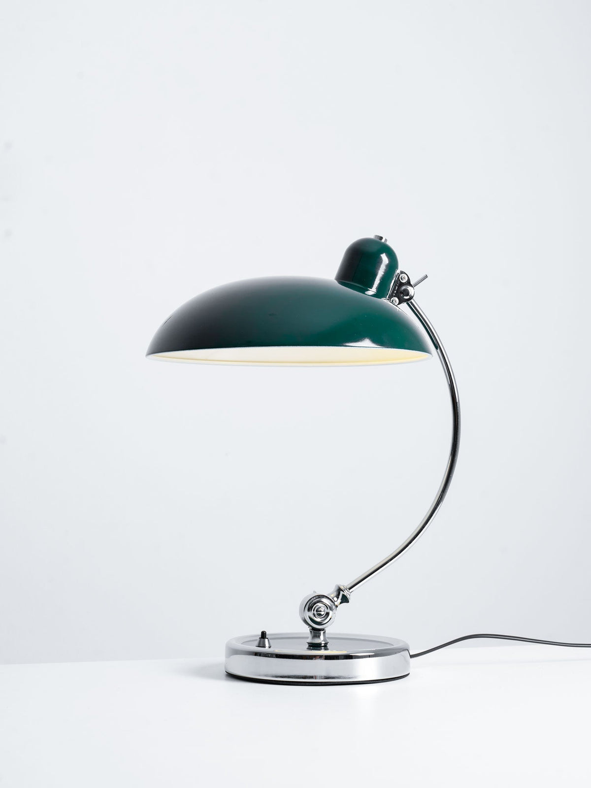 A green Clérachic desk lamp lit on a plain white backdrop, full length.