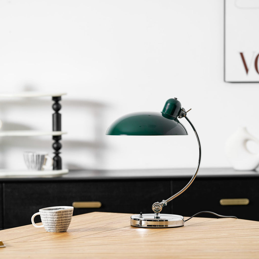 A framed print softens the shelf behind this green Clérachic desk lamp, shown unlit on the wooden table.