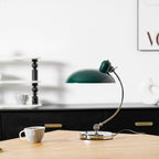 A framed print softens the shelf behind this green Clérachic desk lamp, shown unlit on the wooden table.