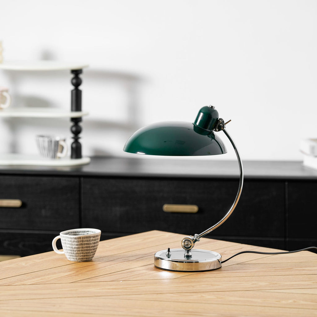 A green Clérachic desk lamp sits on a wooden table beside a ceramic cup, kitchen shelving blurred behind.