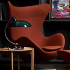 A green Clérachic desk lamp sits on a wooden side table beside a rust-coloured armchair.