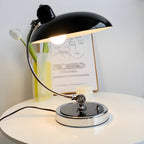 A black Clérachic desk lamp lit beside a tulip stem and a line-art portrait print on a white desk.