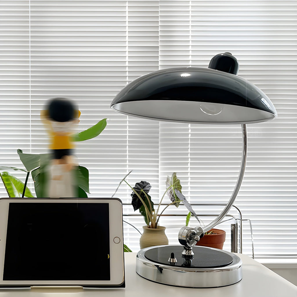 On a desk with potted plants and a tablet, this black Clérachic desk lamp is joined by a blurred toy figure.