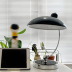 On a desk with potted plants and a tablet, this black Clérachic desk lamp is joined by a blurred toy figure.