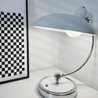 A pale blue-grey Clérachic desk lamp lit beside a black-and-white checked frame on a plain backdrop.