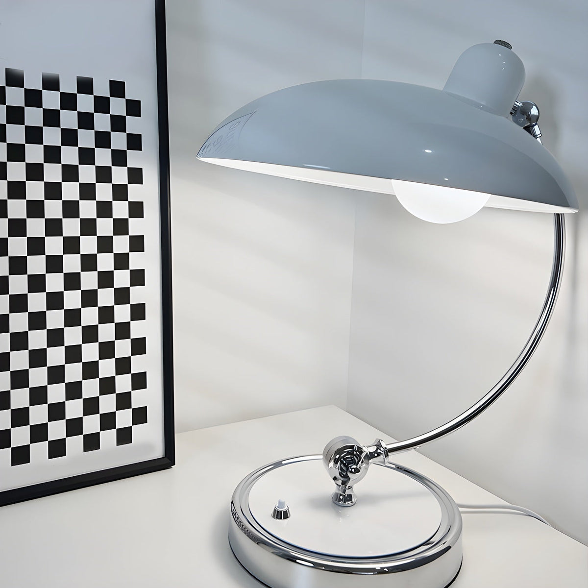 A pale blue-grey Clérachic desk lamp lit beside a black-and-white checked frame on a plain backdrop.