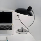 A black Clérachic desk lamp on a white desk beside an open laptop and a wall socket.
