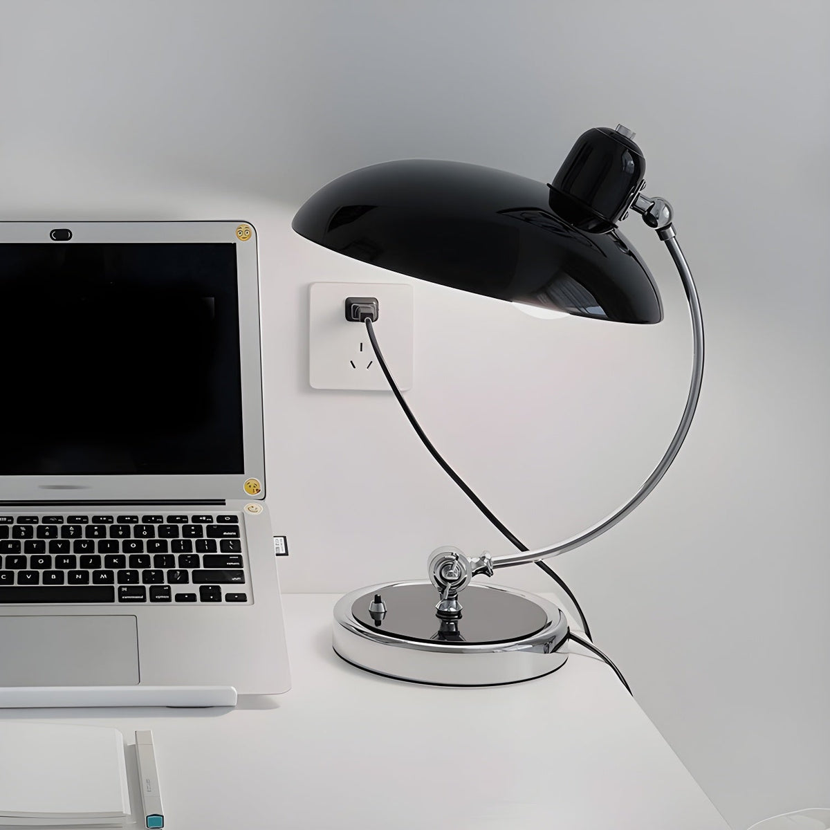A black Clérachic desk lamp on a white desk beside an open laptop and a wall socket.