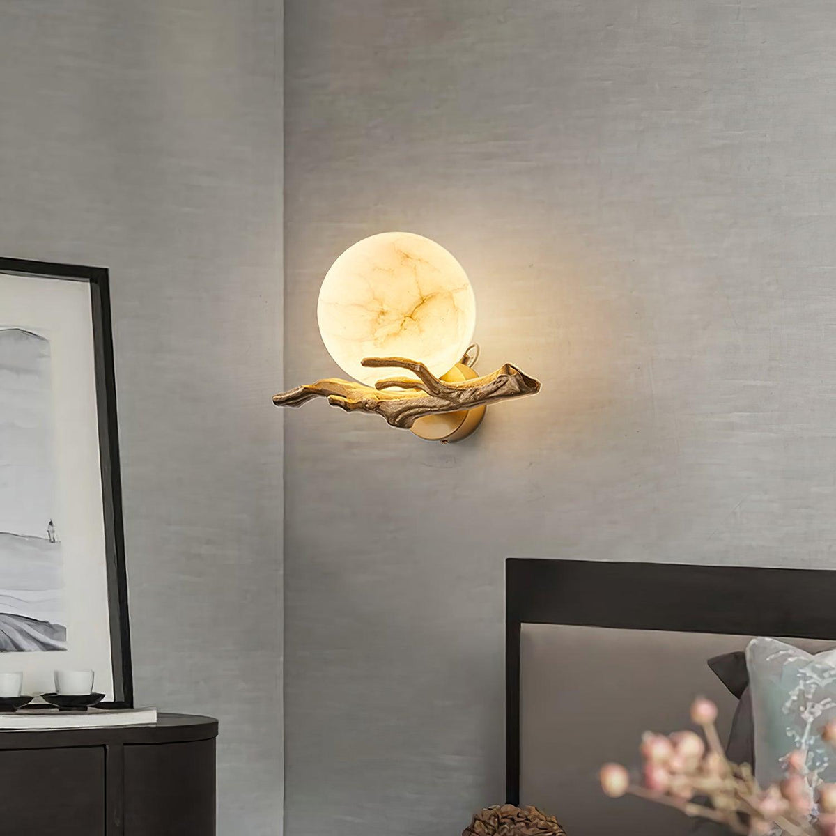 In a bedroom corner, the Clérachic sconce glows above a black dresser dressed with pink flower stems and framed art.