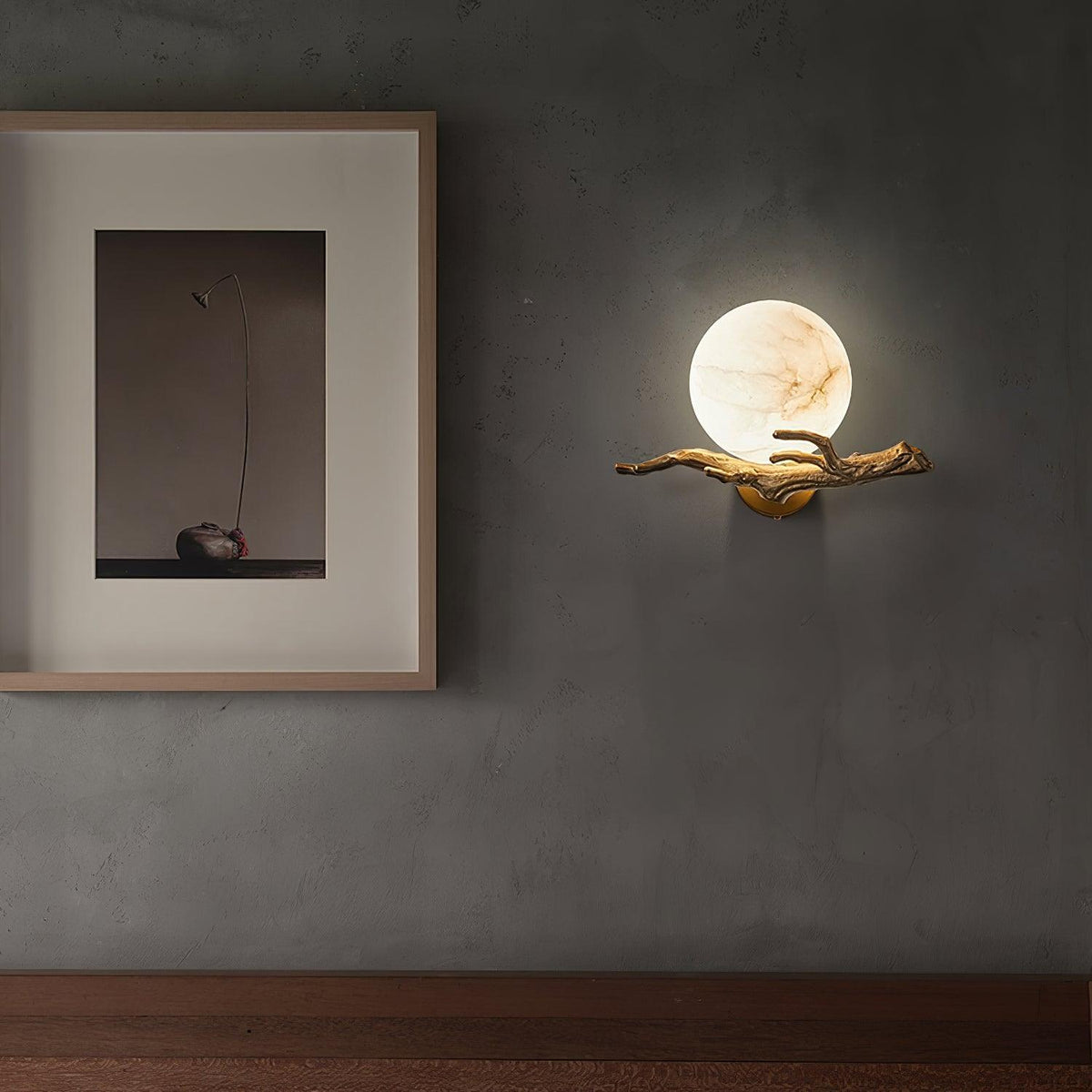 The lit Clérachic sconce hangs on a dark wall above a wood floor, beside a small framed still-life print.