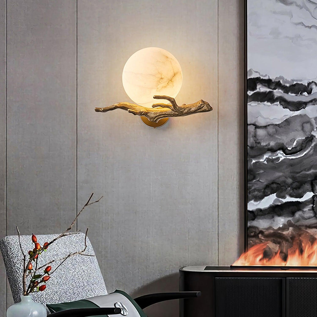 In a living room, the lit Clérachic sconce sits above an armchair, framed abstract art, and a vase of dried branches.