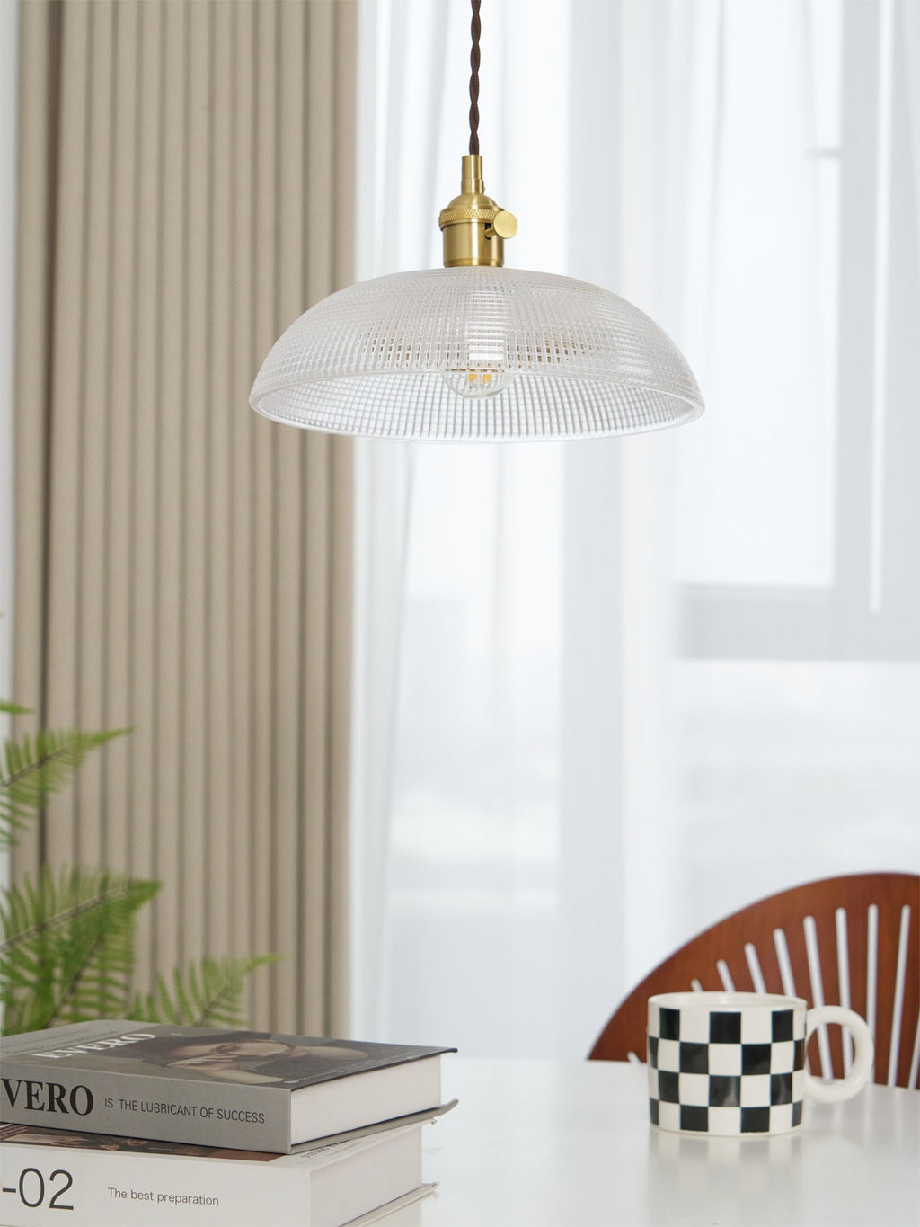 Hanging near a white curtain and a wood chair, this Clérachic pendant sits above books and a chequered mug.